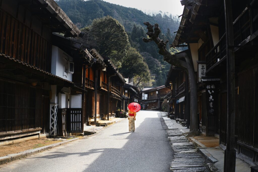 Scenic Japanese village street with a woman in kimono carrying a red umbrella, capturing cultural essence.